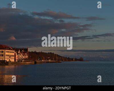 Splendida vista degli edifici e della passeggiata sulla riva del lago di Costanza nella bella luce serale nella stagione invernale con montagne innevate. Foto Stock