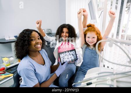 Sorridendo felici bambini, due ragazze multirazziali, dentista in visita, seduto in sedia dentale, gesturing successo con le braccia in su. Giovane donna africana dentista Foto Stock