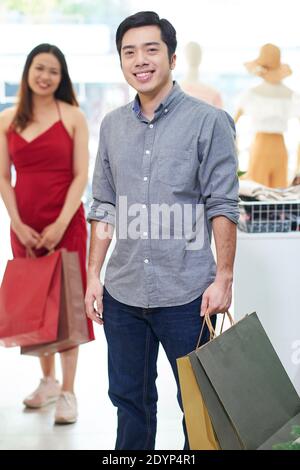 Positivo giovane bell'uomo con borse di carta che guardano la macchina fotografica dopo shopping con la ragazza Foto Stock