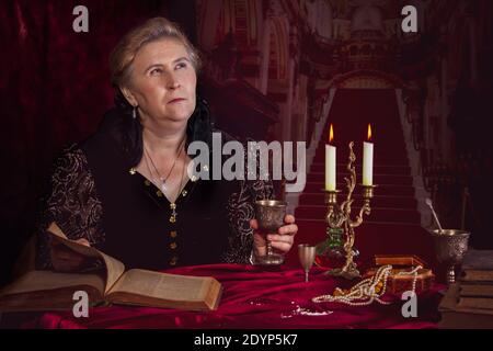 Una donna anziana si trova in un castello medievale con un vecchio libro, candele e una tazza di ferro. Foto Stock