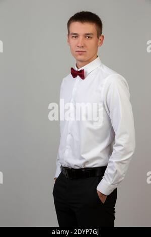 Ritratto classico di un giovane su sfondo grigio. Un ragazzo di circa venti anni in una camicia bianca e cravatta ad arco. Foto Stock