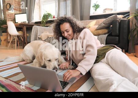 Giovane donna sdraiata sul pavimento con il suo cane e. utilizzo di un computer portatile nel soggiorno Foto Stock