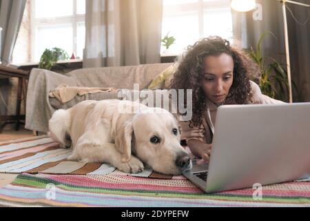 Giovane donna sdraiata sul pavimento con il suo cane e. lavorare online su un computer portatile nella stanza di casa Foto Stock