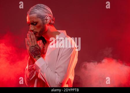 Giovane attore con trucco fase, capelli bianchi in capelli accurati e tatuaggio in piedi davanti alla macchina fotografica e tenendo le mani unite dalla bocca Foto Stock