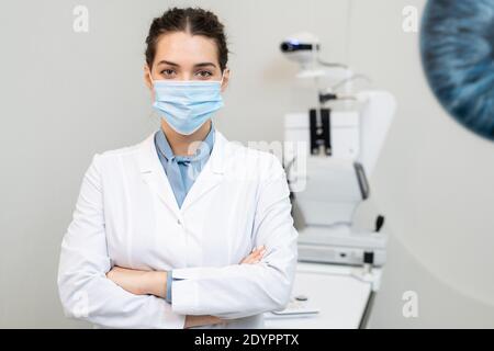 Giovane oftalmologo femminile di brunette in maschera protettiva e passaggio bianco le braccia al petto mentre si trova in piedi contro l'attrezzatura medica Foto Stock