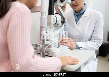 Giovane oftalmologo femminile in whitecoat utilizzando apparecchiature mediche per il controllo vista di una studentessa elementare nelle cliniche contemporanee Foto Stock