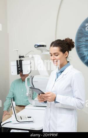 Felice giovane oftalmologo femminile in whitecoat guardando il display tablet sullo sfondo del paziente di sesso maschile anziano che ha la sua vista controllata su Foto Stock