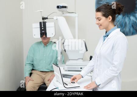 Felice giovane oftalmologista femminile in whitecoat in piedi davanti alle apparecchiature di prova della vista, guardando il display e girando la manopola Foto Stock