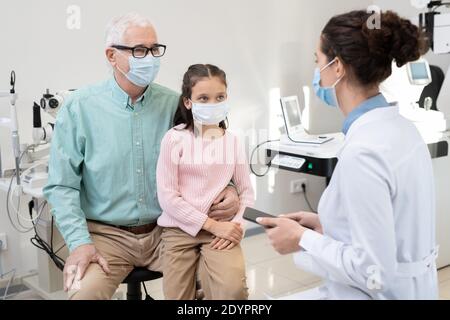 Uomo anziano e sua nipote in maschere protettive seduti dentro fronte di giovane oftalmologo femminile durante la consultazione medica in cliniche Foto Stock