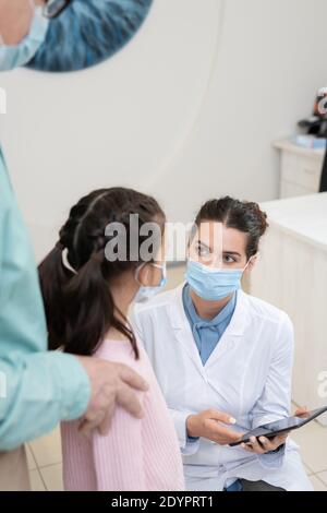 Giovane oftalmologista femminile in maschera protettiva e whitecoat utilizzando compresse mentre dà il consiglio medico alla scolastica ed al suo nonno Foto Stock