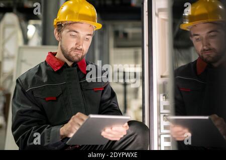 Giovane ingegnere serio in abbigliamento da lavoro e hardhat scorrendo attraverso tecnico dati in tavoletta digitale di una delle macchine industriali Foto Stock