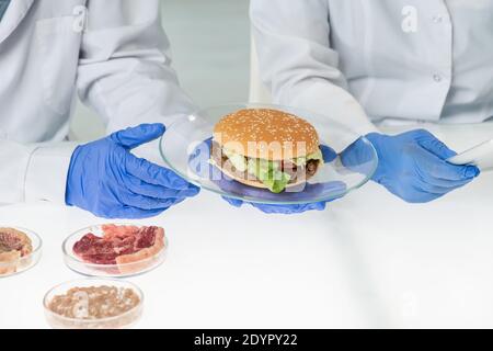 Mani guelse del lavoratore della piastra di tenuta per il controllo della qualità degli alimenti con hamburger appetitoso contenente carne vegetale sopra tavola in laboratorio Foto Stock