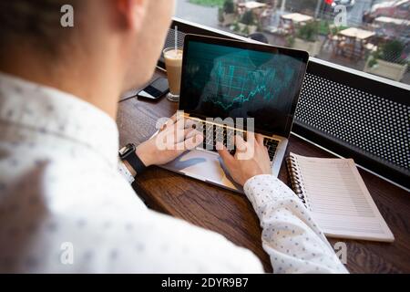 Uomo freelance che lavora su un computer portatile nella caffetteria. Grafici finanziari sullo schermo. Concetto di investimenti di mercato. L'uomo di successo lavora a distanza. Finanziario Foto Stock