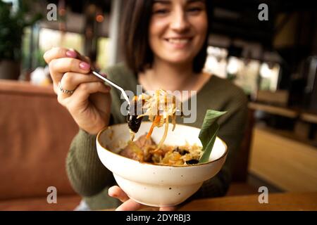 Donna felice offuscata che mostra il suo pasto gustoso e sorridente. Concetto di cibo asiatico. Ristorante o caffetteria sullo sfondo. La giovane donna bruna gode del favorito Foto Stock