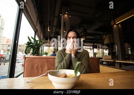 Una giovane donna malata con naso che cola venne in un bar. Posto pubblico sul pankground. La donna bruna soffia il naso. Isolatevi se vi ammalate Foto Stock