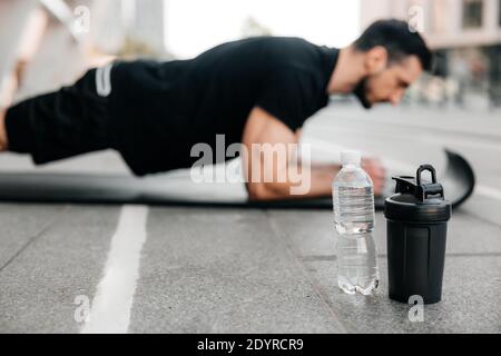 Uomo concentrato che fa la posizione della tavola mentre si esercita sul tappeto di yoga nero. Bottiglie d'acqua che si trovano vicino. Uomo offuscato in nero sport esercizio Foto Stock