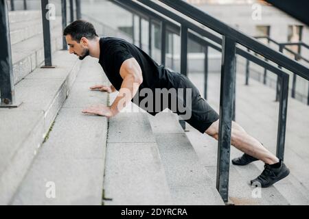 Giovane uomo ben costruito che si spinge verso l'alto dalle scale. Vista laterale. Uomo atletico con capelli neri che indossa abbigliamento sportivo nero e sneakers che si allenano in strada Foto Stock