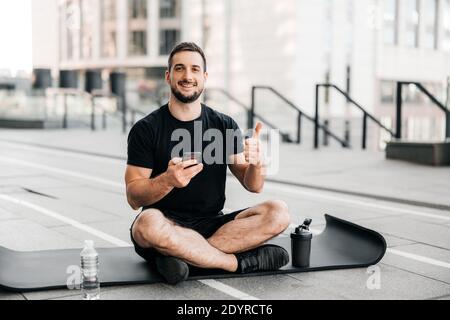 Uomo che riposa dopo l'esercizio fisico all'aperto con smartphone in mano e che si fa avanti. Bottiglia d'acqua sportiva su tappetino nero per yoga. Uomo sportivo dopo la pratica Foto Stock