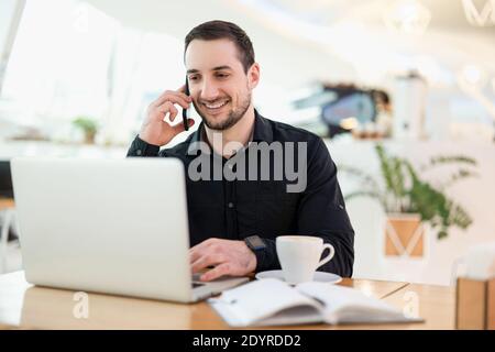 Lavoratore maschile remoto di successo. Giovane freelance felice di una telefonata. Caffetteria sullo sfondo. Programmatore freelance maschile che lavora in un caffè Foto Stock