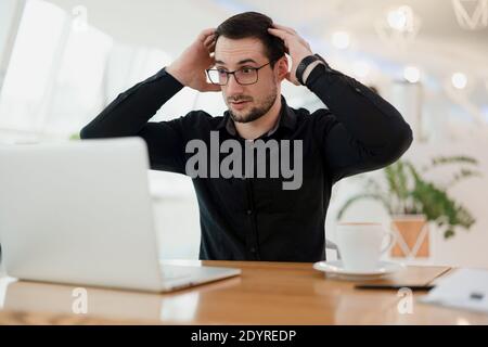 Che cosa sta accadendo uomo intelligente scioccato in occhiali guardando sul suo laptop e tenendo la testa per le mani. Errore nel programma. Il programmatore maschile è scioccato Foto Stock