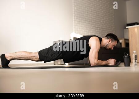 Corsi di formazione IT a casa. Giovane uomo sicuro che indossa abiti sportivi neri e che fa la posizione della tavola mentre si esercita sul pavimento in un loft leggero interno Foto Stock