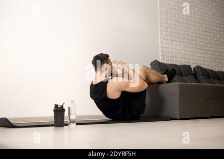 Pressa di pompaggio dell'uomo atletico con le gambe sul divano. Allenamento duro. Concetto di stile di vita sano. Un moderno soggiorno di colore bianco sullo sfondo. Uomo forte che si escerca Foto Stock