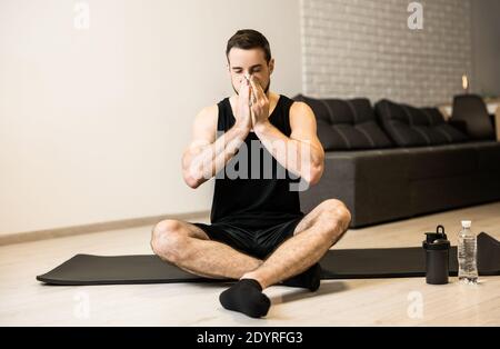 Uomo malato con naso che cola seduto sul tappeto nero yoga. Concetto di soggiorno a casa. Il giovane bruna gli soffia il naso dopo l'allenamento. Praticare sport a casa se Foto Stock