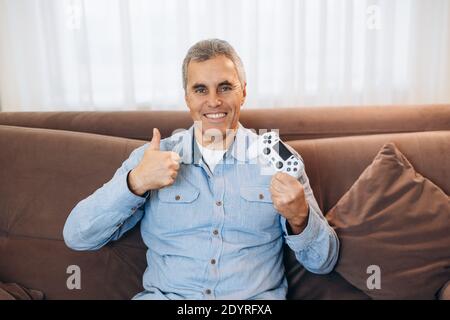 Vinci in un videogioco Middle Aged Man che mostra il suo controller di gioco, che si mette in evidenza, sorridendo con i denti e guardando la fotocamera. L'uomo allegro è felice a causa di Foto Stock