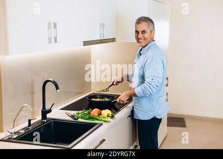 Uomo di mezza età felice di cucinare qualcosa di gustoso e sano per la sua famiglia. In piedi in stufa, stufò le verdure in una padella, sorridendo e guardando Foto Stock