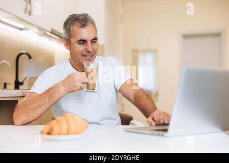 Uomo maturo che gli gustava il cappuccino e il croissant yummy. Concetto di routine mattutina. Un freelance maschile sta gustando la sua dolce colazione, guardando il computer portatile Foto Stock