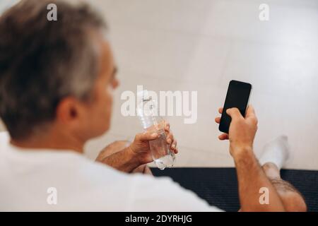 Uomo anziano che usa lo smartphone e tiene una bottiglia d'acqua in plastica. Sopra la spalla colpo. Uomo in t-shirt bianca seduto sul tappeto nero yoga e riposante Foto Stock