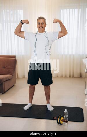 Orgoglioso uomo maturo che mostra i suoi muscoli. Concetto di allenamento duro. Fit uomo anziano mostrando bicipiti e sorridendo. Soggiorno luminoso sullo sfondo. Bell'uomo Foto Stock
