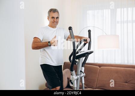L'uomo felice maturo che si accerca sull'addestratore ellittico e che thumbing in su. Allenati a casa. Allenamento cardio con cross-trainer. Soggiorno bianco su Foto Stock