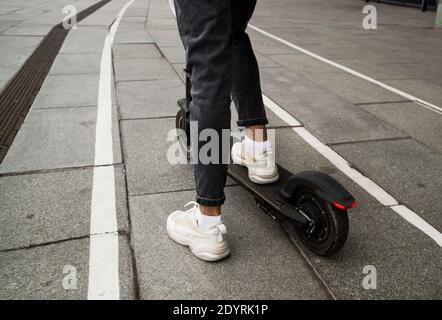 Gambe di anonimo ragazzo che cavalcano moderno scooter elettrico su strada asfaltata su sfondo sfocato della strada della città. Concetto di viaggio veloce. Giovane uomo alla moda Foto Stock
