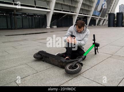 L'uomo cadde dallo scooter che gli teneva il ginocchio e sentiva dolore. Scooter elettrico verde su asfalto. Un uomo elegante con cappuccio grigio si siede a terra e. Foto Stock