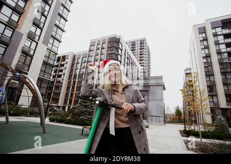 Una donna sorridente viaggia su uno scooter elettrico noleggiato in tutta la città. Lei gioca con il cappello rosso di babbo natale e con il suo laugthing. Giovane allegra donna bionda dentro Foto Stock