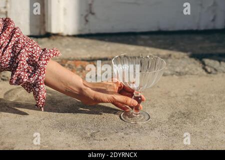 Primo piano di donne mano in abito floreale che tiene vuoto bicchiere di champagne alla luce del sole. Concetto di Capodanno, matrimonio o festa di compleanno con bevanda. Selez Foto Stock