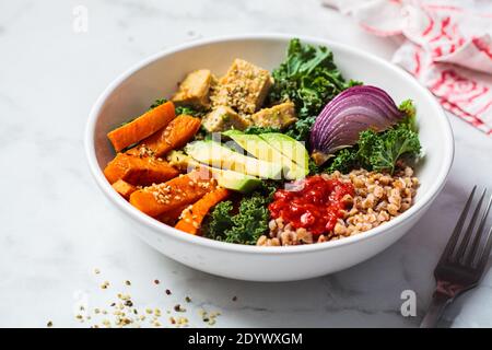 Verdure cotte al forno, avocado, tofu e ciotola di buddha di grano saraceno. Insalata vegana con kale, patate dolci al forno, tofu, grano saraceno e avocado in un arco bianco Foto Stock