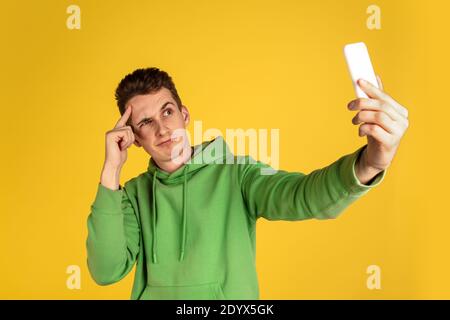 Selfie. Ritratto di un giovane caucasico su sfondo giallo dello studio. Bel modello maschile in outfit verde con cellulare. Concetto di emozioni umane, espressione facciale, vendite, pubblicità, bellezza. CopySpace. Foto Stock