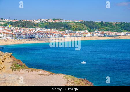 Vista aerea della barca bianca in acque turchesi azzurre Di Oceano Atlantico e spiaggia sabbiosa costa Praia da Nazare città con piccole figure di persone Foto Stock