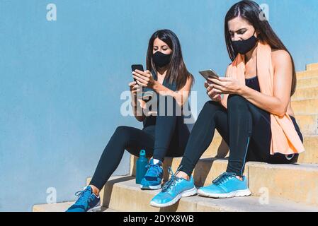 due giovani donne vestono abiti sportivi e indossano maschere nere seduta su gradini facendo una pausa e controllando i loro smartphone. Concetto di sport Foto Stock