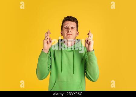 Dita incrociate. Ritratto di un giovane caucasico su sfondo giallo dello studio. Bel modello maschile in outfit verde gesturing. Concetto di emozioni umane, espressione facciale, vendite, annuncio, gioventù. CopySpace. Foto Stock