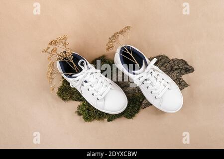 Concetto etico di scarpe vegan. Un paio di sneakers bianche con fiori secchi su corteccia di alberi e muschio, fondo di carta da mestiere beige neutro. Foto Stock