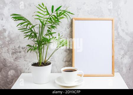 Telaio verticale in legno con cartoncino bianco, tazza di caffè e fiore verde esotico di palma in vaso su tavolo su fondo grigio parete in cemento. Templ. Mockup Foto Stock