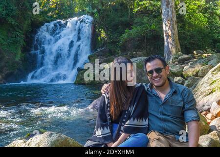 Giovane coppia ispanica felice seduta su una pietra circondata da belle scene di natura Foto Stock