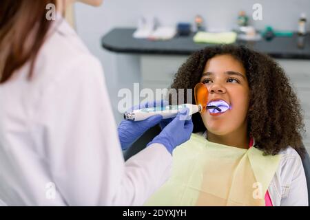 Vista posteriore del dentista donna che indossa uniforme bianca, che fornisce il ripristino del dente e il riempimento con polimerizzazione lampada UV per il suo piccolo paziente Foto Stock