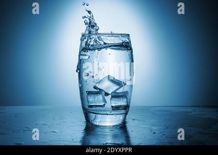Un bicchiere d'acqua con cubetti di ghiaccio. H2O spruzzato in un recipiente trasparente. Tonalità blu. Foto Stock