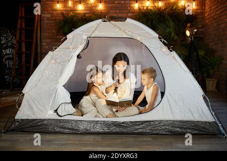 Madre legge un libro di fiabe per i suoi figli mentre si siede in una tenda di notte. Mamma, figlio e figlia che leggono un libro con una torcia elettrica nella loro h Foto Stock