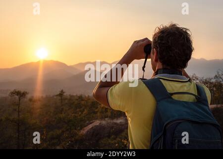 Un giovane uomo con uno zaino sulla schiena guarda attraverso il binocolo al tramonto alle sagome del montagne Foto Stock