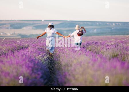 Ragazzo e ragazza dalla schiena in un campo di lavanda e abiti rustici. Camminano con una chitarra e tengono le mani Foto Stock